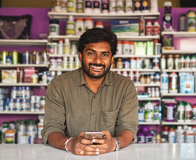 An agri-input dealer holds his mobile and leans smilingly over his counter. In the background, there is a full shelf of various agricultural products.