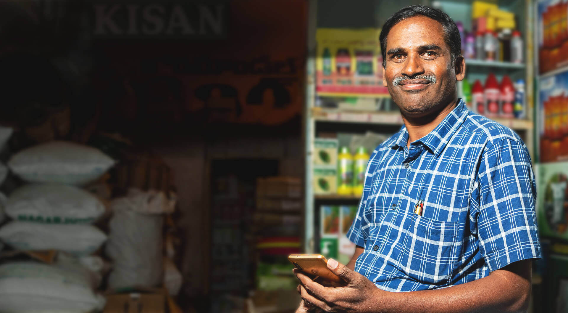 प्लेड शर्ट में कृषि उत्पादों का एक विक्रेता स्मार्टफ़ोन हाथ में लिए अपनी दुकान में आत्मविश्वास के साथ खड़ा है। उसके चेहरे पर संतोष झलक रहा है।